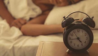 A woman lies in bed looking at the alarm clock.